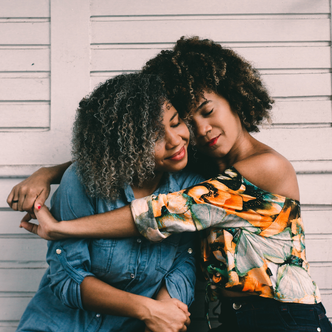 Two women in an embrace in front of a white wall.