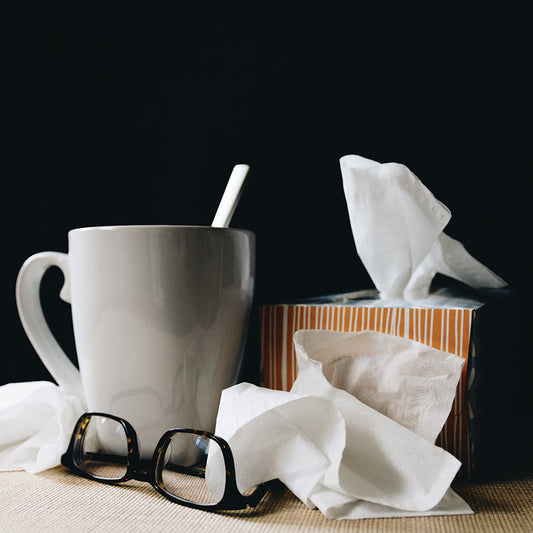 A white drinking mug and a box of facial tissues with used tissues and a pair of glasses.