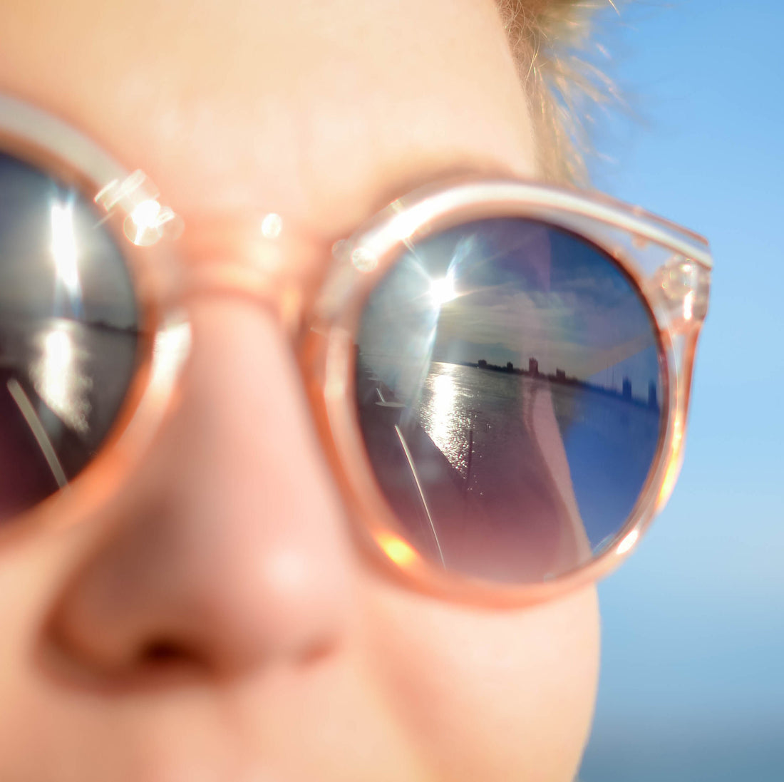 Closeup of sunglasses on a person’s face showing the reflection of a city skyline, bright sun, and a clear blue sky.