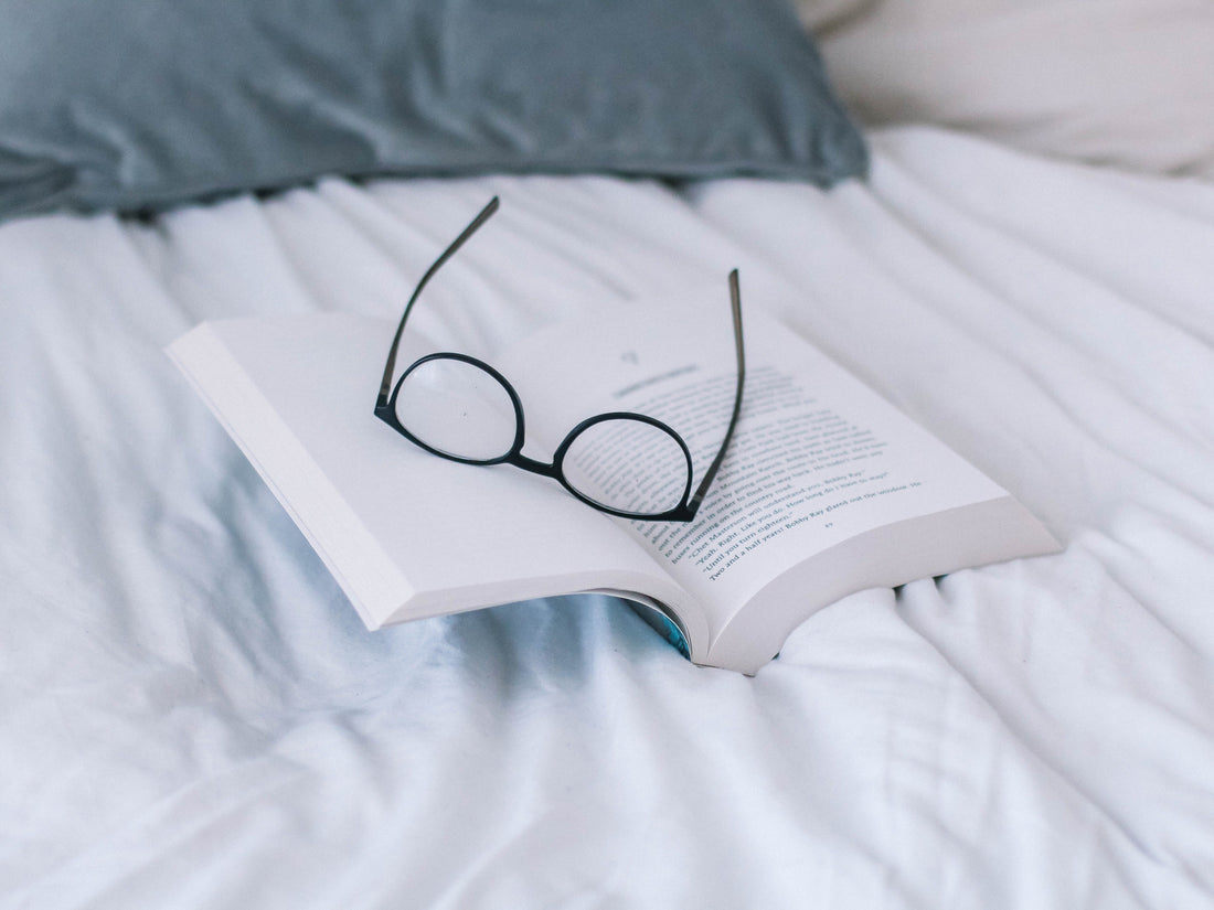 A pair of glasses sitting on top of an open book that is sitting on top of a bed with white covers and a grey pillow