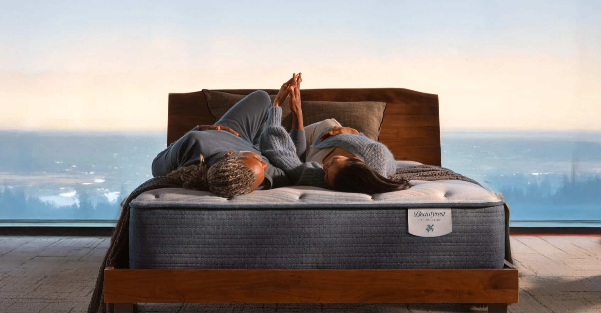 Corner of a Beautyrest Harmony mattress with a cream blanket draped on it and sitting in a light brown wood bed frame. .  