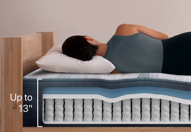 A woman sleeps comfortably on her side on a multi-layered mattress with a cutaway view, showcasing its foam and coil construction. The mattress thickness is labeled 'Up to 13' inches.