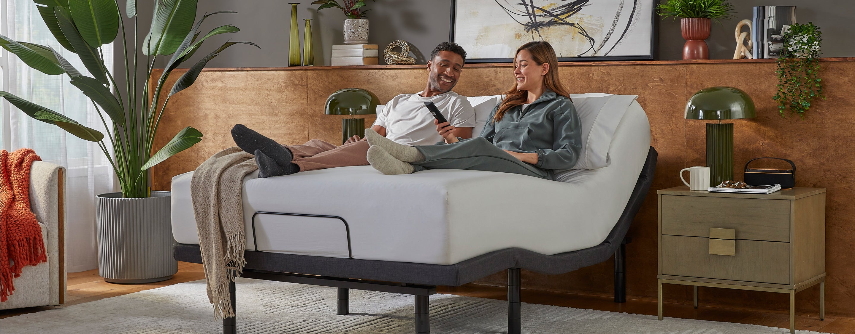 Man and woman sitting on a white Serta mattress, sitting on baselogic base with her holding the remote to the base