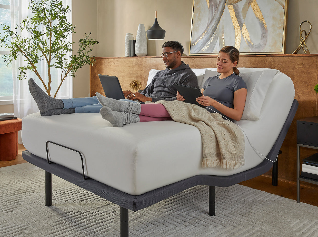 Man and woman using their laptop and tablet devices sitting on a white Serta mattress on a baselogic bedframe