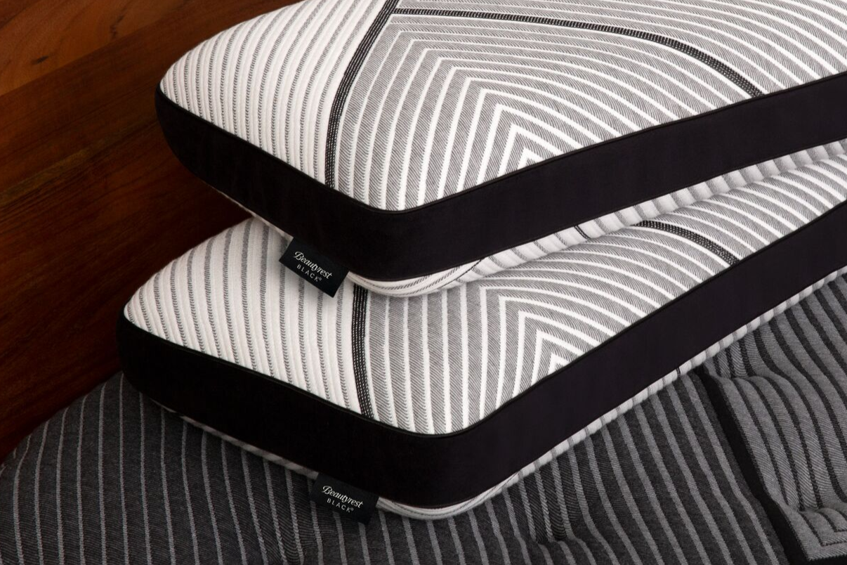 Two stacked pillows with a modern black-and-white geometric design, featuring black edges and a 'Beautyrest Black' label, resting on a textured gray mattress.