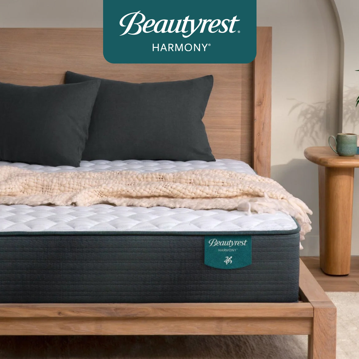 A Beautyrest Harmony mattress with a white quilted top on a wooden bed frame, styled with dark green pillows and a cozy cream blanket, designed for support, pressure relief, and a lifted feel.