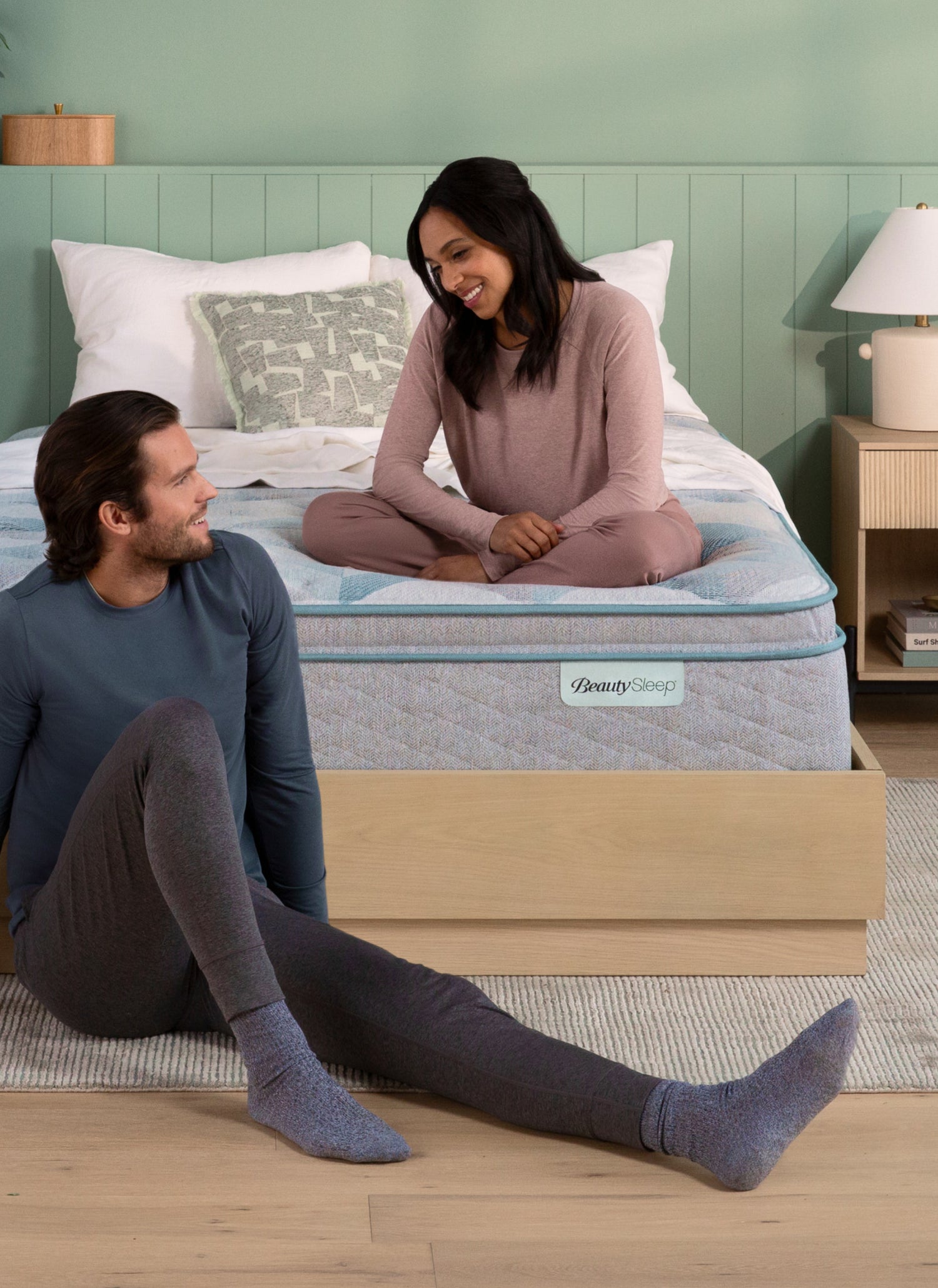 A couple in cozy loungewear sitting and smiling near a BeautySleep mattress on a wooden bed frame in a softly styled bedroom