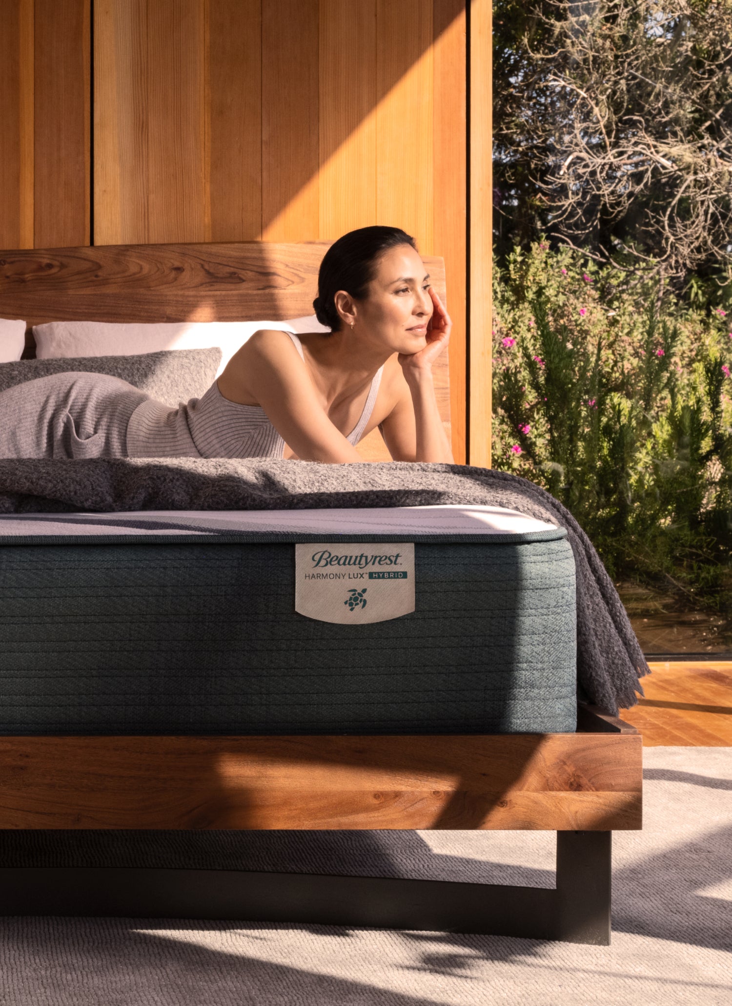 A woman lounging on a Beautyrest Harmony Lux Hybrid mattress on a wooden bed frame, with sunlight streaming in and a garden visible through a large window