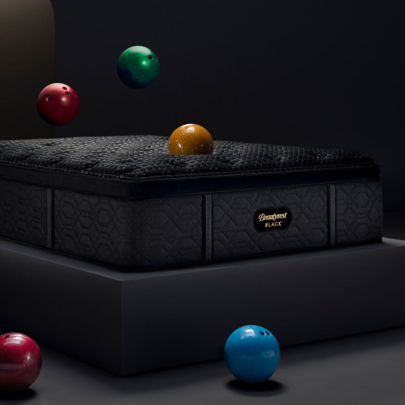 Image of a red, green and yellow bowling ball falling onto an Beautyrest Black mattress sitting on a grey platform with a pink and blue bowling ball on the floor in front of it.