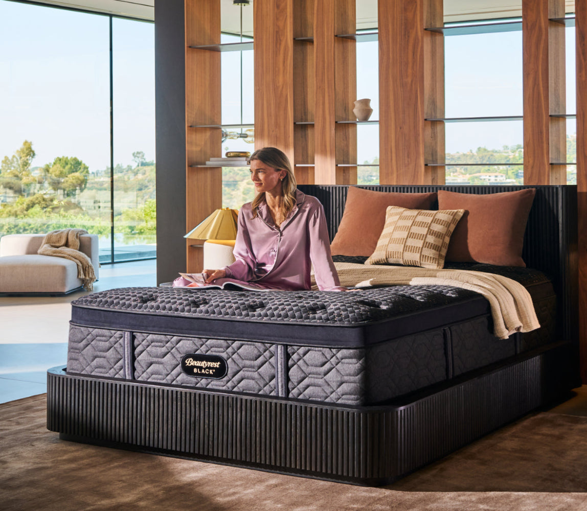 Woman sitting up, reading a magazine, on a Beautyrest Black mattress with 3 pillows and a blanket on it.