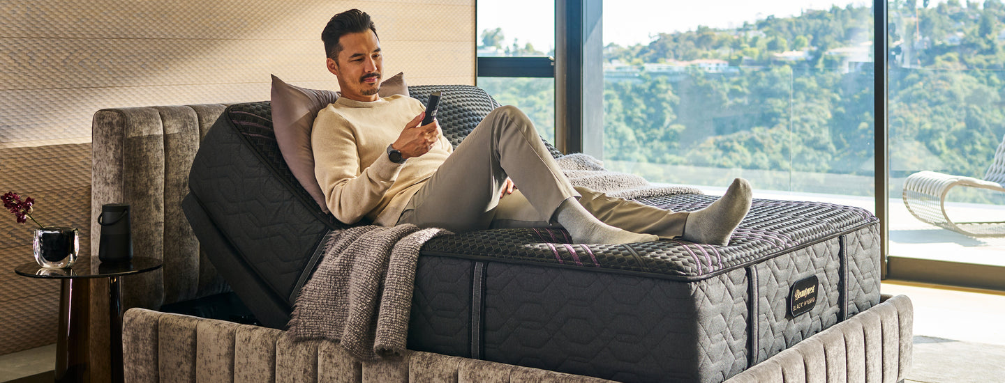 Man sitting on a Beautyrest adjustable bed looking at the remote for it with big windows showing lots of trees in the background.