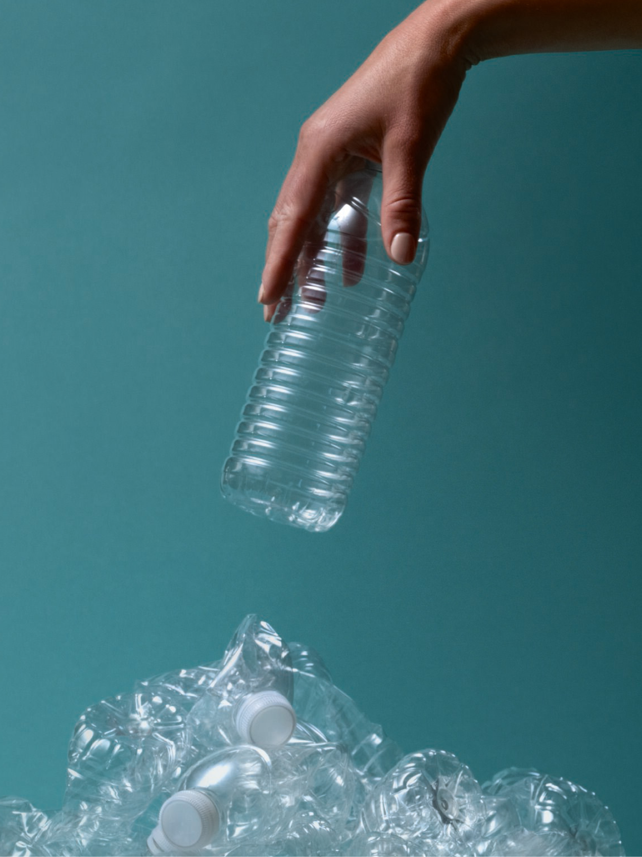 "A hand dropping a clear plastic bottle onto a pile of crushed plastic bottles, symbolizing recycling and environmental consciousness against a teal background.